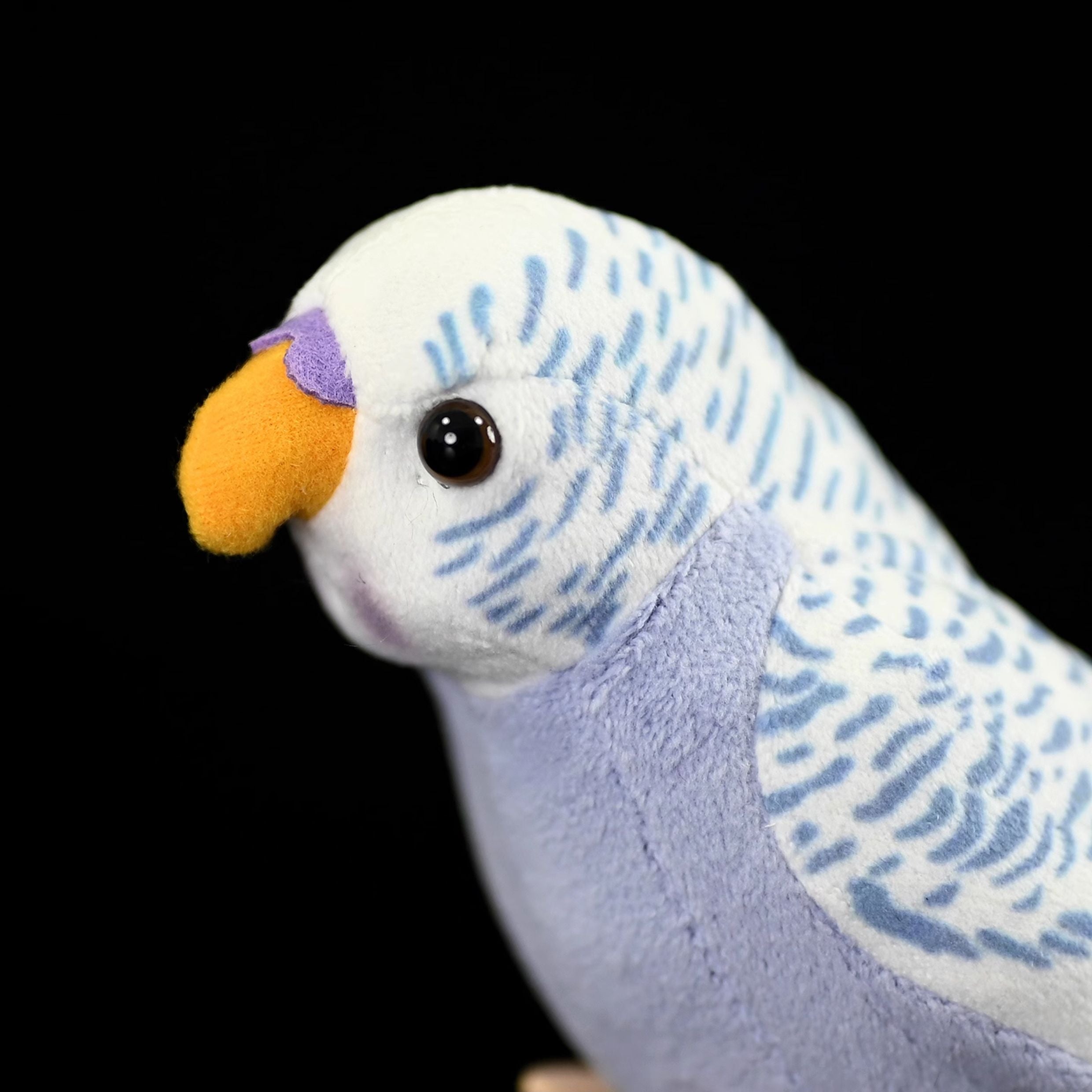Close-up of a violet budgerigar plush toy's head, featuring bright eyes and an orange beak