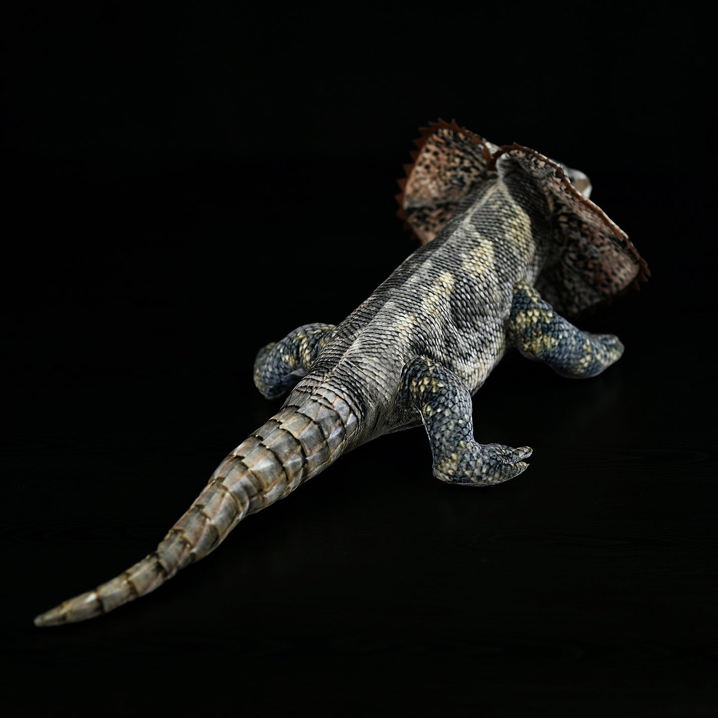 Rear view of a frilled lizard plush, emphasizing its long, textured tail and detailed reptile-like design